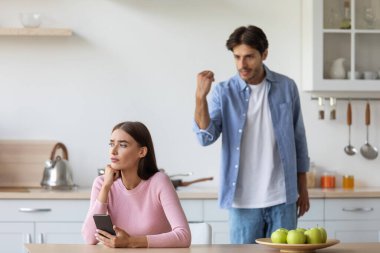 Kızgın üzgün, üzgün genç adam mutfakta akıllı telefonuyla karısına bağırıyor.