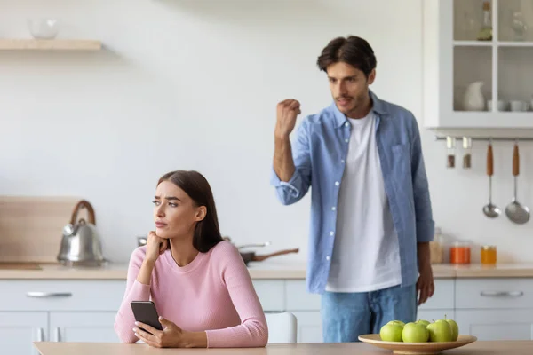 Enojado molesto joven triste gritándole a la esposa con teléfono inteligente en el interior de la cocina — Foto de Stock