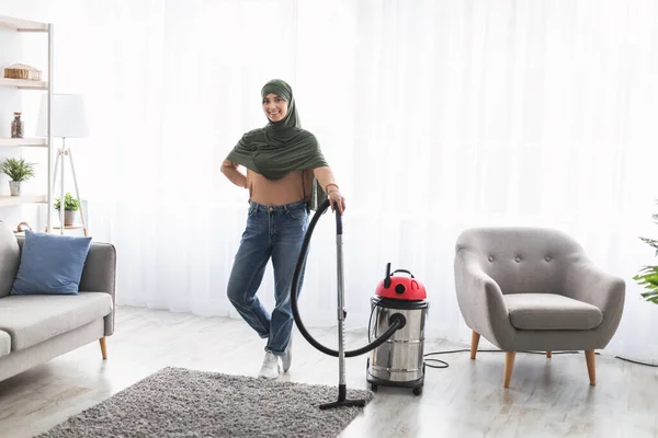 Mulher muçulmana alegre tapete de limpeza com aspirador de pó, posando — Fotografia de Stock