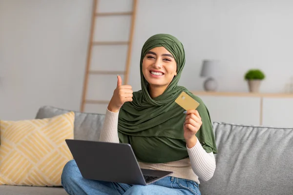 Feliz joven árabe en hijab gesto pulgar hacia arriba, la celebración de la tarjeta de crédito, compras en la tienda web en el ordenador portátil en casa — Foto de Stock