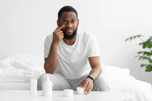 Σοβαρή χιλιετή μαύρο γενειοφόρος άνθρωπος εφαρμόζει κρέμα στο πρόσωπο, κάθεται στο κρεβάτι στο υπνοδωμάτιο με βάζα των καλλυντικών — Φωτογραφία Αρχείου