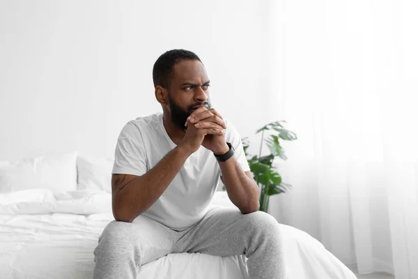 Δυστυχισμένος σκεπτικός νεαρός μαύρος γενειοφόρος άνδρας κάθεται στο κρεβάτι στο εσωτερικό του υπνοδωματίου σε λευκό φόντο τοίχου — Φωτογραφία Αρχείου
