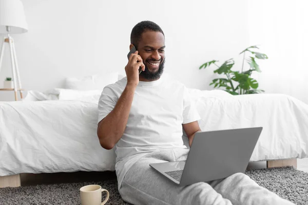 Χαμογελώντας millennial μαύρο γενειοφόρο αρσενικό κάθεται στο κρεβάτι εργάζονται σε φορητό υπολογιστή μιλάμε στο τηλέφωνο με τον πελάτη — Φωτογραφία Αρχείου