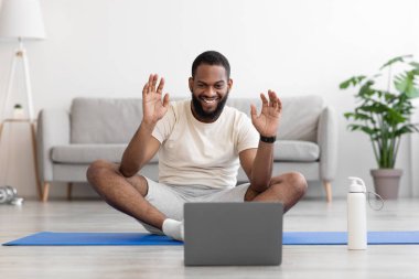 Gülümseyen genç siyah erkek oturma odasında paspasın üzerine oturmuş şişeyle bilgisayara bakıyor.