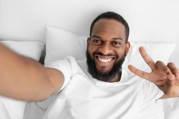 Χαμογελώντας νεαρός μαύρος γενειοφόρος άντρας ξαπλώνει στο λευκό κρεβάτι στην κρεβατοκάμαρα, βγάζει σέλφι και δείχνει το σημάδι ειρήνης. — Φωτογραφία Αρχείου