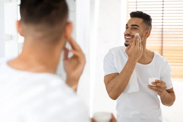 Cuidado de belleza masculino. Hombre árabe joven guapo aplicando crema hidratante para la cara — Foto de Stock