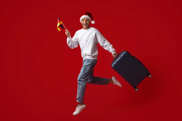 Radosny młody facet w kapeluszu Santa skacząc z niemiecką flagą i walizką — Zdjęcie stockowe