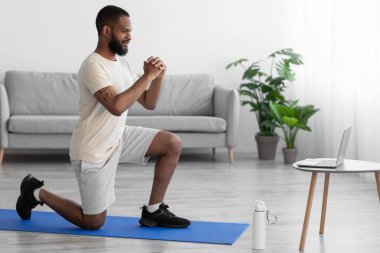 Gülümseyen bin yıllık Afro-Amerikan sakallı adam odanın içinde paspasın üzerinde bacak egzersizi yapıyor.