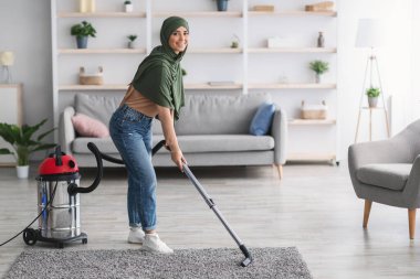 Halıyı elektrikli süpürgeyle temizleyen neşeli Müslüman kadın.