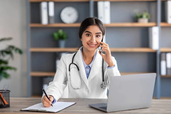 Χαμογελώντας χιλιετή εκ των υστέρων θεραπεύτρια γιατρός σε λευκό παλτό συμβουλεύεται ασθενή, μιλώντας τηλεφωνικώς στο γραφείο κλινική — Φωτογραφία Αρχείου