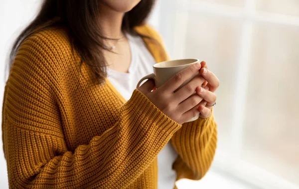 Vista cortada da mulher árabe aquecendo as mãos na xícara de café quente perto da janela em casa, espaço vazio — Fotografia de Stock