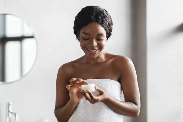 Mujer negra sonriente cubierta de toalla usando producto para el cuidado del cuerpo —  Fotos de Stock