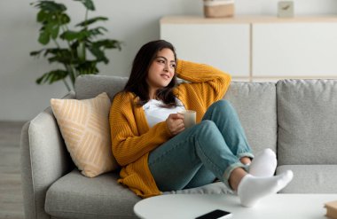 Huzurlu genç Arap kadın bir fincan kahveyle kanepede dinleniyor, yan bakıyor ve hayal kuruyor.