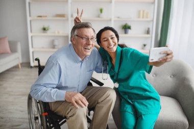 Happy senior manin wheelchair taking selfie with lovely young nurse at home clipart