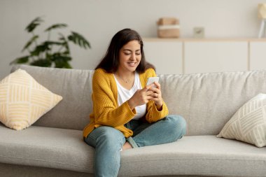 Genç Arap bayan evdeki kanepede otururken akıllı telefon kullanıyor, arkadaşlarıyla mesajlaşıyor ya da yeni uygulamalara göz atıyor.