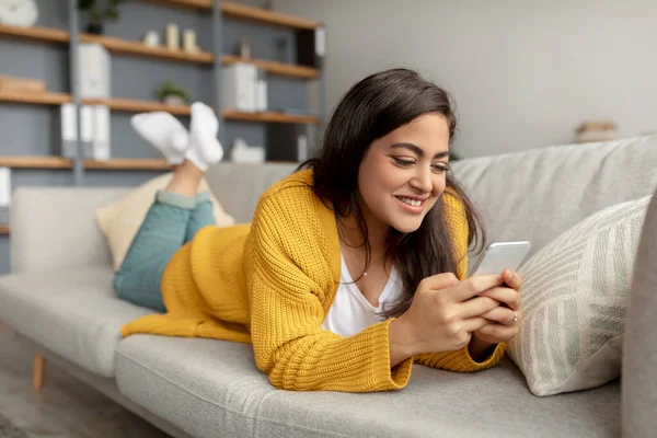 Affascinante signora araba sdraiata sul divano con smartphone, controllare i social media, chattare online a casa, spazio libero — Foto Stock