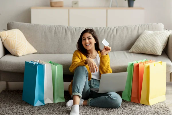 Heyecanlı Arap kadın büyük satışlardan dolayı mutlu hissediyor, dizüstü bilgisayar kullanıyor ve kredi kartını gösteriyor, hediye çantalarıyla yerde oturuyor. — Stok fotoğraf