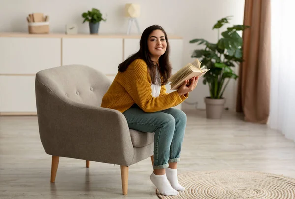 Glad ung lugn muslimsk kvinna sitter på bekväm fåtölj och läsa pappersbok, njuter av morgonen och ler — Stockfoto