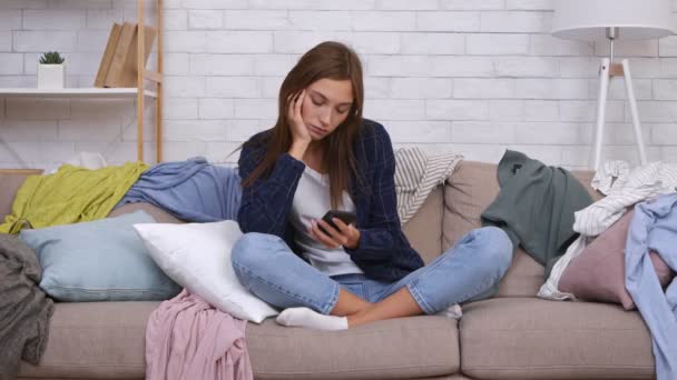 Joven perezosa mujer indiferente sentado entre la ropa dispersa en el sofá y navegar por la web en el teléfono inteligente, ignorando el desorden — Vídeo de stock