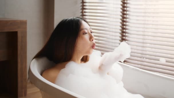 Όμορφη ασιατική γυναίκα φυσώντας αφρό, ενώ λαμβάνοντας ζεστό μπάνιο στο σπίτι — Αρχείο Βίντεο