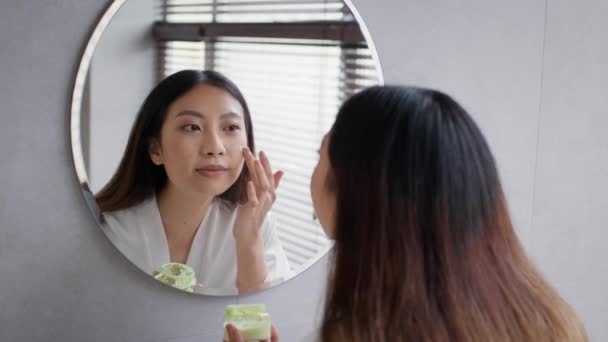 Cuidado de la piel. Hermosa mujer asiática mirando al espejo y aplicando crema hidratante para la cara — Vídeos de Stock