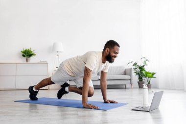 Beyaz spor giyimli mutlu siyah erkek bacak egzersizi yapıyor ve dizüstü bilgisayarda video izliyor.