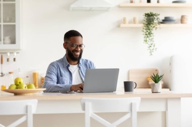 Gülümseyen bin yıllık Afro-Amerikan sakallı adam minimalist mutfak iç mekanında dizüstü bilgisayarla çalışıyor.