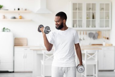 Gülümseyen bin yıllık yakışıklı sakallı Afrikalı beyaz elbiseli Amerikalı erkek sporcu minimalist mutfakta dambılları kaldırıyor.