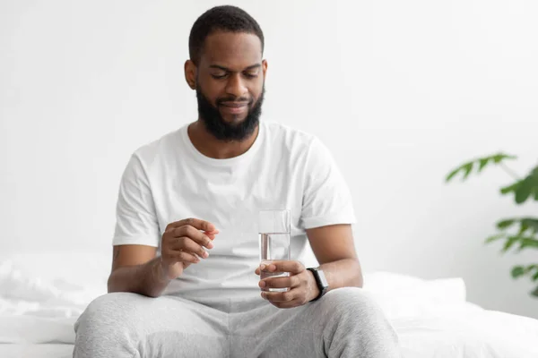 Grave joven negro barbudo macho sentado en la cama en el dormitorio, mantenga el vaso de agua y la tableta — Foto de Stock