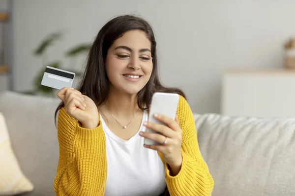 Online payment concept. Cheerful arab lady holding credit card and smartphone, sitting on sofa, making internet purchase — Stock Photo, Image