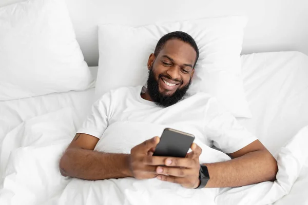 Χαμογελώντας νεαρός μαύρος γενειοφόρος τύπος βρίσκεται στο λευκό κρεβάτι στην κρεβατοκάμαρα, ελέγχει την εφαρμογή και τα κοινωνικά δίκτυα — Φωτογραφία Αρχείου