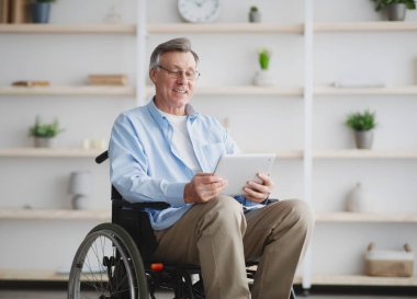 Tekerlekli sandalyedeki neşeli yaşlı adam tablet bilgisayar kullanıyor, internette geziniyor.