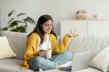 Genç Arap kadın evdeki video görüşmesi için dizüstü bilgisayar kullanıyor, kanepede otururken web kamerasına konuşuyor ve el kol hareketi yapıyor.