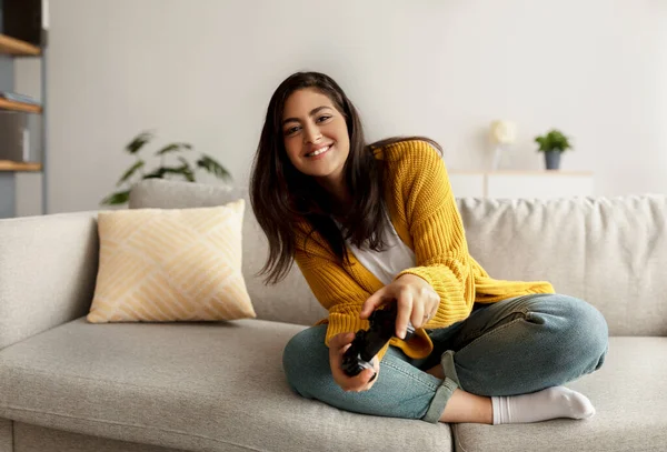 Evde kalıp eğlenmek için. Ortadoğulu neşeli kadın online video oyunları oynamak için joystick kullanıyor, kanepede oturuyor.