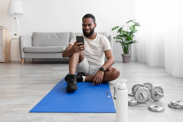 Sonriente millennial africano americano barbudo sentarse en la estera en el suelo en el interior de la habitación con pesas, botella de agua — Foto de Stock