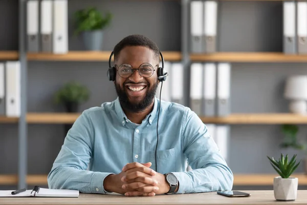 Glimlachende duizendjarige Afrikaanse bebaarde man in bril en headset werken op laptop en kijken naar camera — Stockfoto