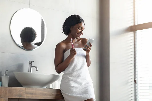 화장실에서 이를닦으면서 스마트폰을 사용하는 흑인 여성 — 스톡 사진