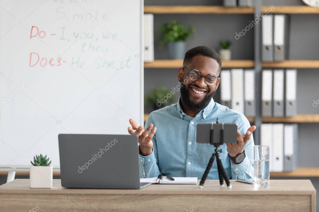 Happy young african american bearded guy teacher in glasses works at laptop and looks at webcam