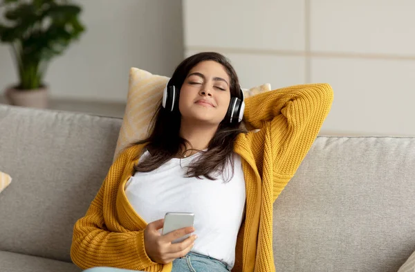 若いリラックスしたアラブの女性のワイヤレスヘッドフォンでリラックスソファで、自宅で音楽を聴き、フリースペース — ストック写真