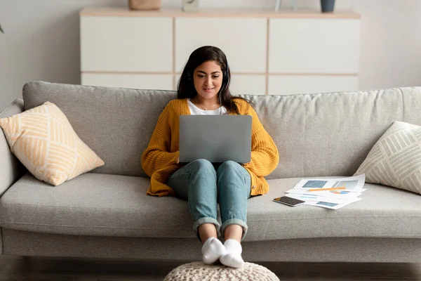 Young arab female freelancer using laptop wearing wireless earphones and typing on keyboard, sitting on sofa at home — Stock Photo, Image