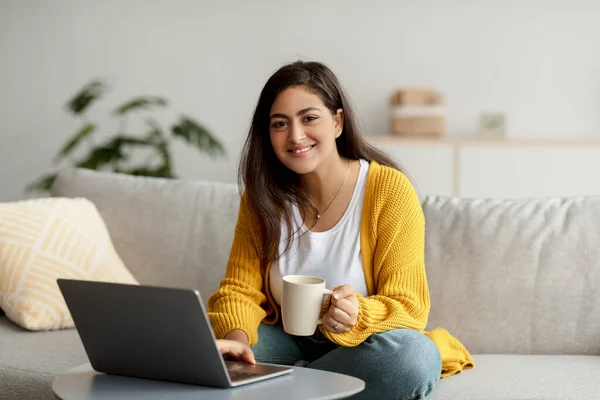 Portret van een vrolijke arabische vrouwelijke freelancer die koffie drinkt en laptop gebruikt, thuis online werkt, ruimte kopieert — Stockfoto