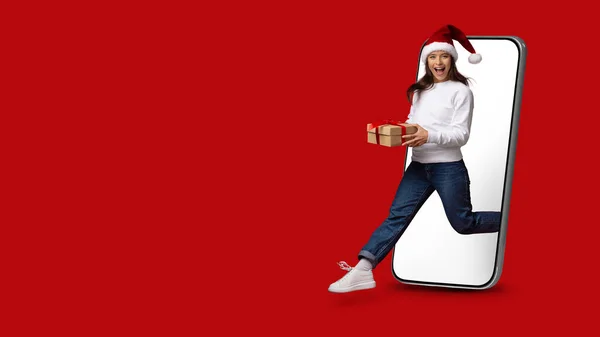 Lady in Santa Hat Holding κουτί δώρου πηδώντας έξω από το μεγάλο Smartphone — Φωτογραφία Αρχείου