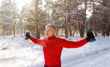 Kış parkında, panorama 'da dambıllarla egzersiz yapan aktif kıdemli bir adam. Soğuk havada antrenman.