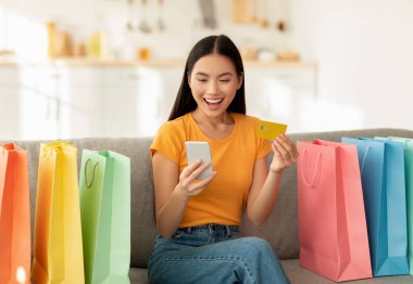 Evden alış veriş. Kredi kartı ve akıllı telefonlu aşırı neşeli Asyalı kadın online siparişini onaylıyor.