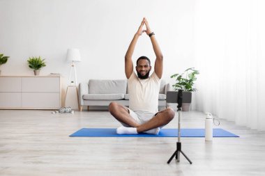 Nilüfer pozisyonunda beyaz spor giyimli, sakallı, Afro-Amerikan bir adam. Telefon kamerasındaki videoyu kaydediyor.