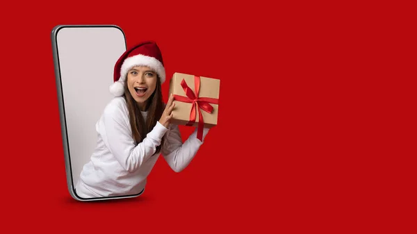 Xmas Surprise. Lady In Santa Hat Holding Gift Peeking Out Of Smartphone — Stock Photo, Image