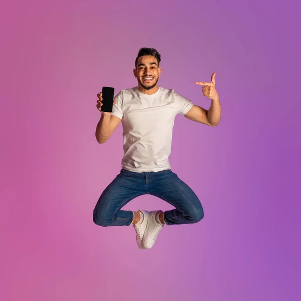 Funky pemuda Arab melompat dan menunjukkan smartphone modern dengan layar kosong di lampu neon, mockup — Stok Foto