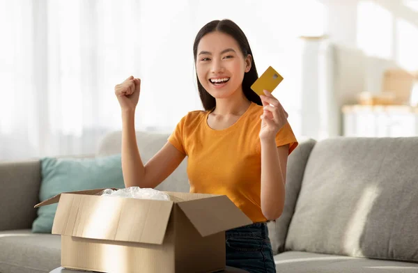 Šťastná asijská žena přijímající balíček, zaťaté pěsti a ukazující kreditní kartu, spokojená se svým nákupem — Stock fotografie
