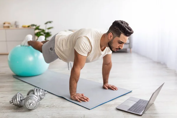 Fuerte chico árabe de pie en tablón delante de la computadora portátil, utilizando la pelota de fitness, ejercitando a tutorial de deportes en línea en casa —  Fotos de Stock