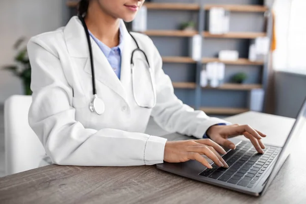 Cortado jovem médico mulher hindu em casaco branco com estetoscópio digitando no computador no interior do escritório — Fotografia de Stock